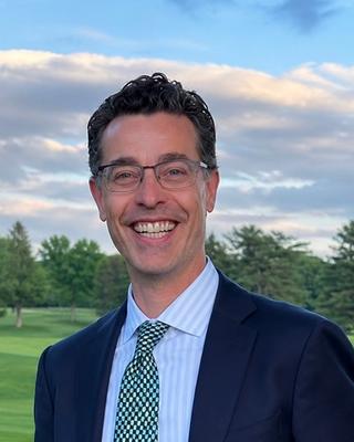 A headshot of Michael Balboni wearing a blue suit and and green tie with a green lawn with trees and blue sky with clouds in the background. 