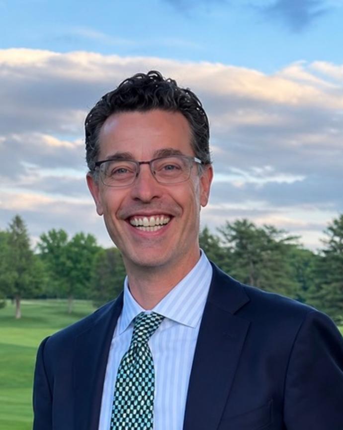 A headshot of Michael Balboni wearing a blue suit and and green tie with a green lawn with trees and blue sky with clouds in the background. 
